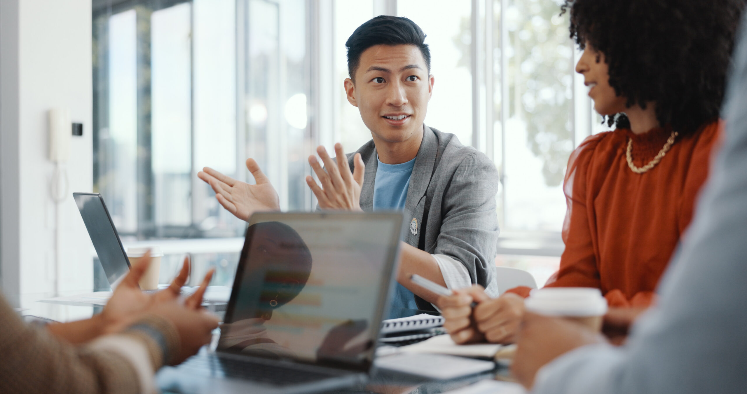 Business professionals in an office meeting discuss strategy around a table with laptops and tablets.
