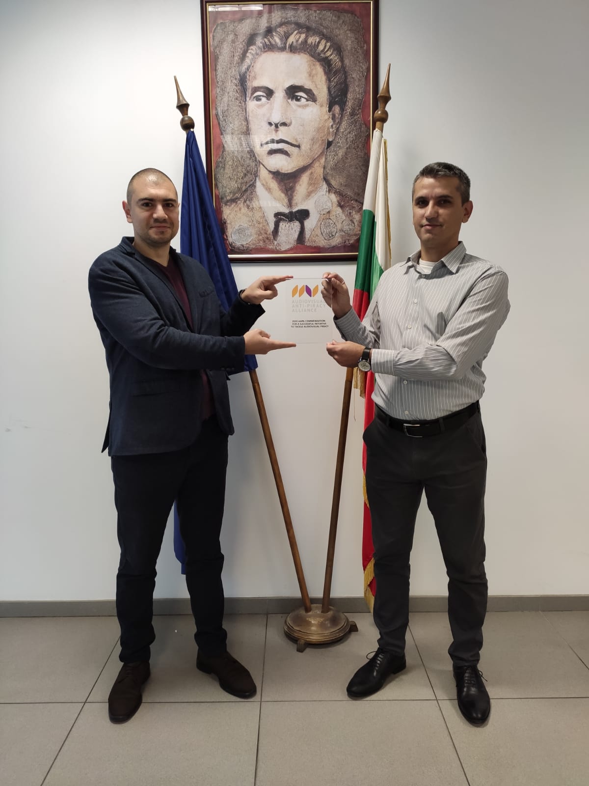 Deux hommes présentent un document entre deux drapeaux, devant un portrait encadré.