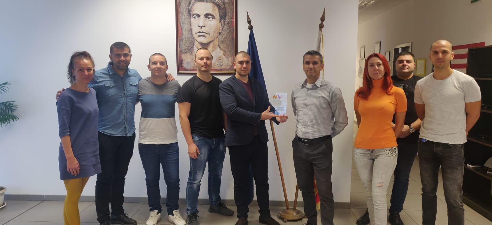 Ten individuals stand together indoors before a portrait and flag, one holding a certificate.
