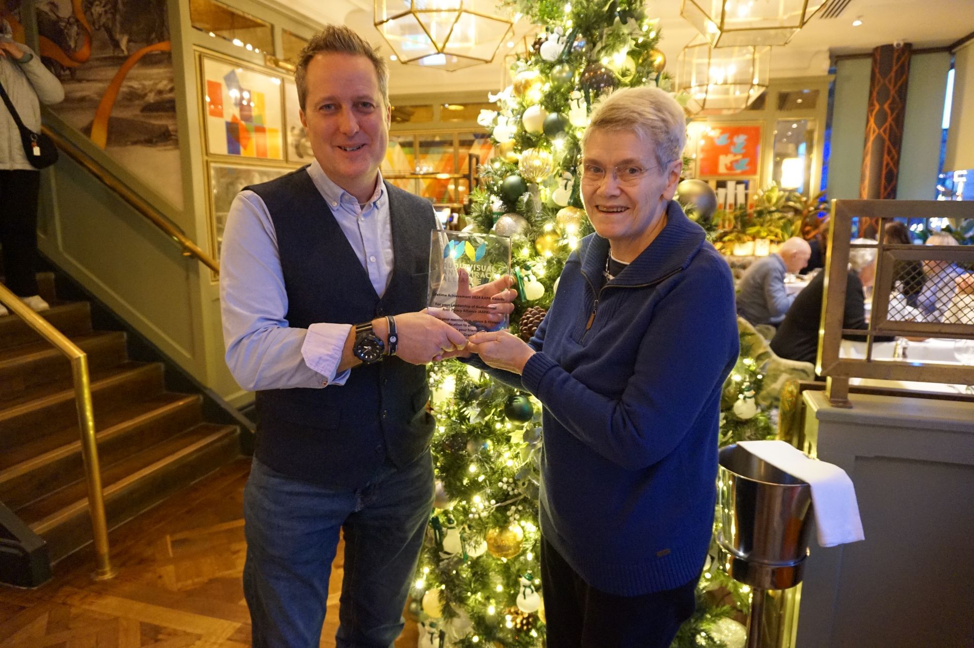 Chris Prior Sheila Cassells — deux personnes échangent un prix devant une décoration de Noël dans un intérieur élégant.