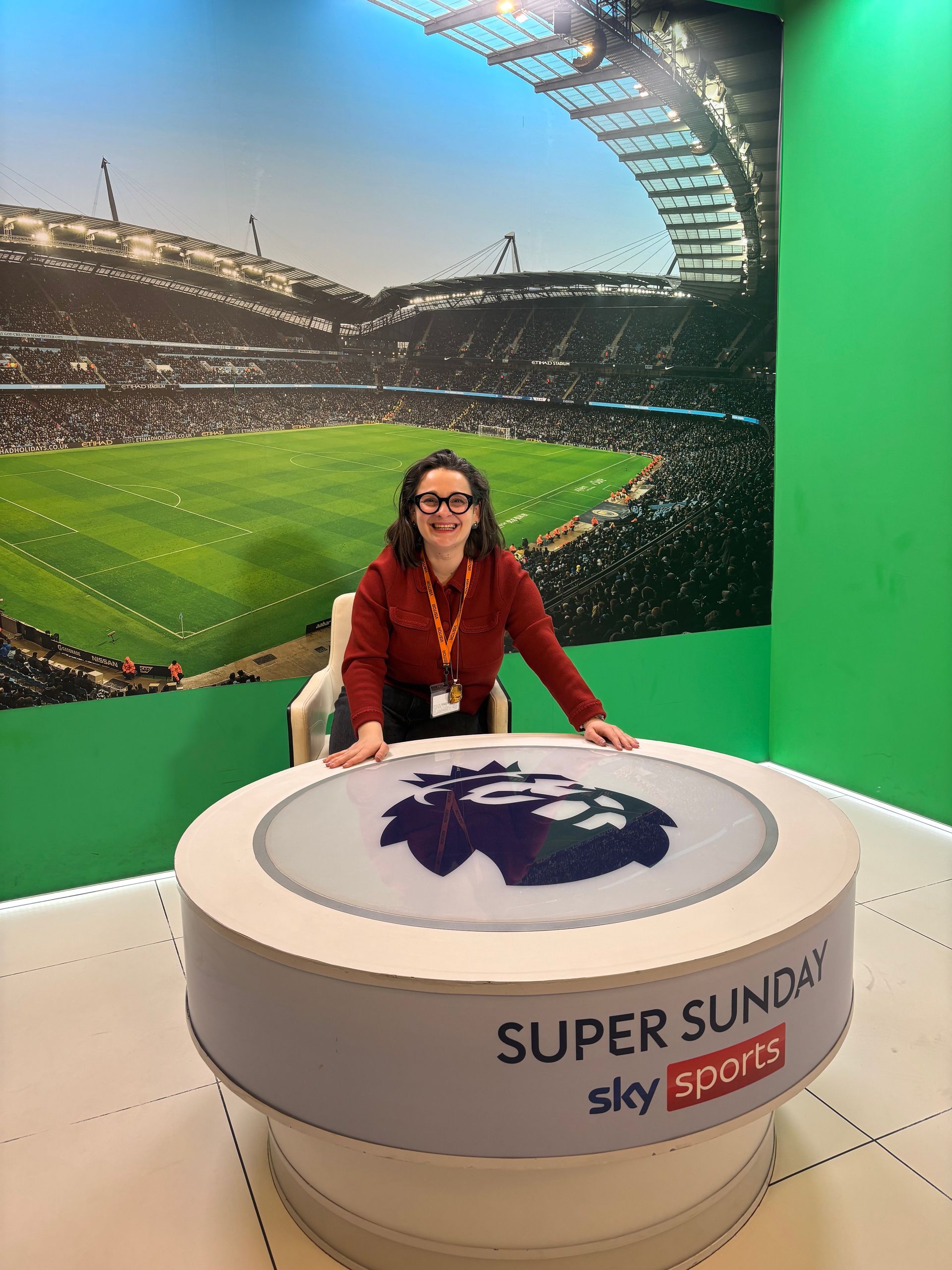 Une femme portant un badge se tient devant une table de studio de télévision pour le football, avec un stade en arrière-plan.