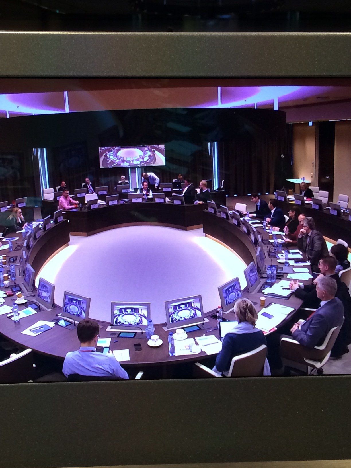 Réunion institutionnelle dans une salle de conférence en fer à cheval avec participants autour d'une table circulaire, écrans de surveillance et…