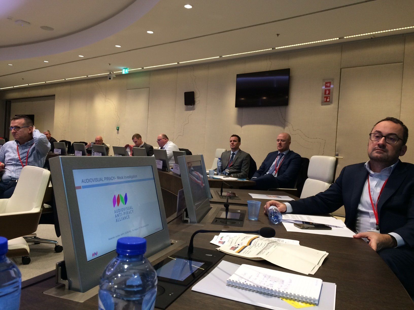 Participants seated at a conference table during an AAPA meeting on audiovisual piracy, with a monitor displaying the AAPA logo and agenda.