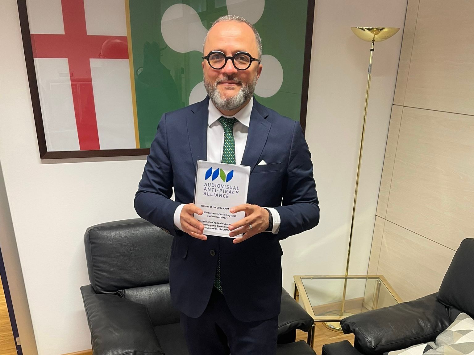Landscape Massimiliano Capitanio Agcom — a man in a dark suit and green tie holds an AAPA award plaque whilst standing in an office setting with…