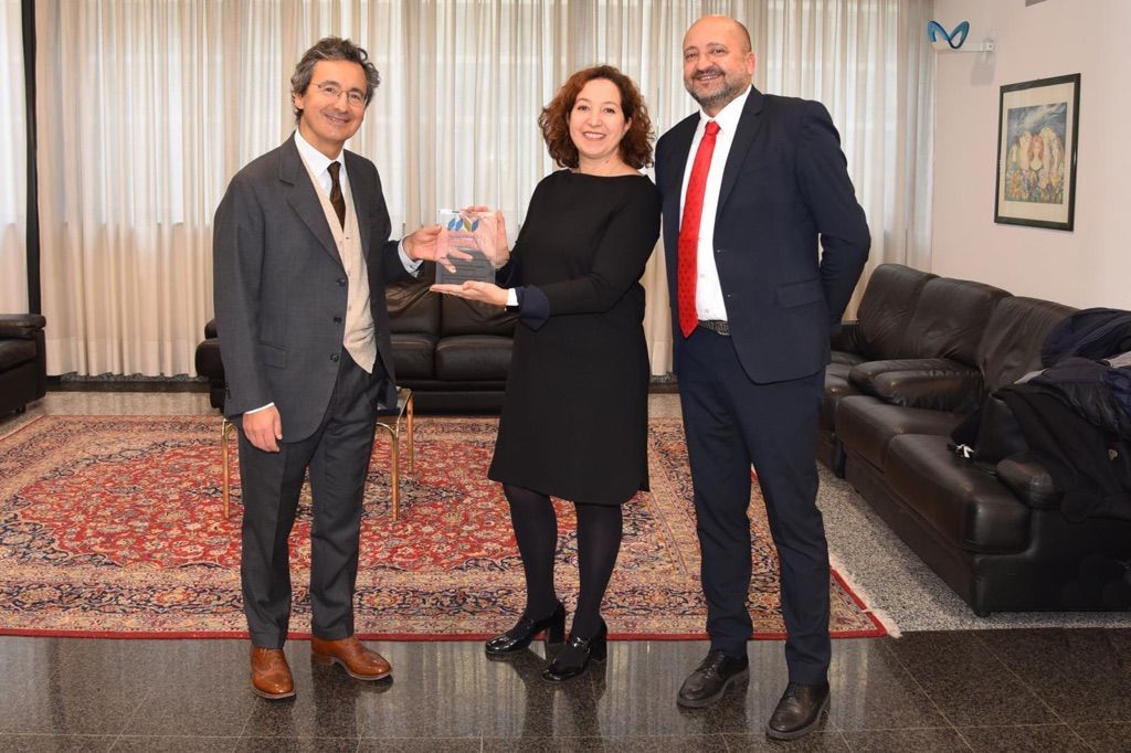 Three professionals exchange a document in a formal office setting with modern furnishings and Italian institutional décor.
