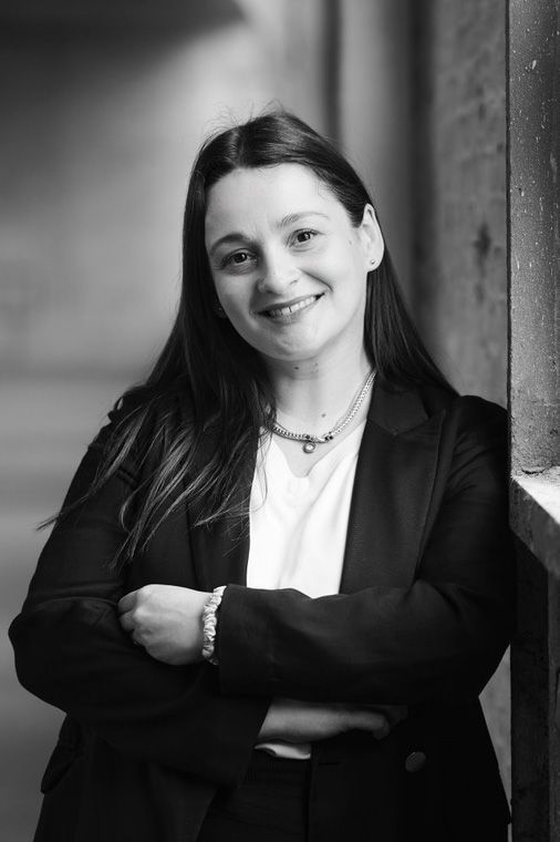 Femme en costume noir et chemise blanche, souriante, appuyée contre un pilier dans un cadre urbain en noir et blanc.