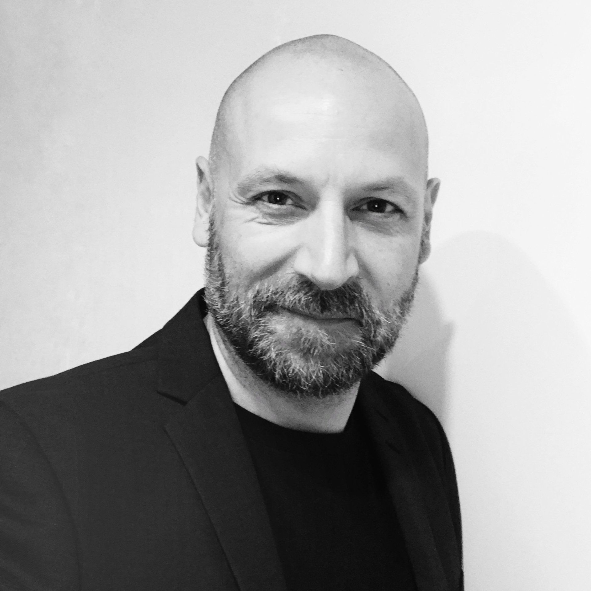 Portrait en noir et blanc d'un homme chauve avec une barbe, vêtu d'une veste noire sur un pull à col rond.