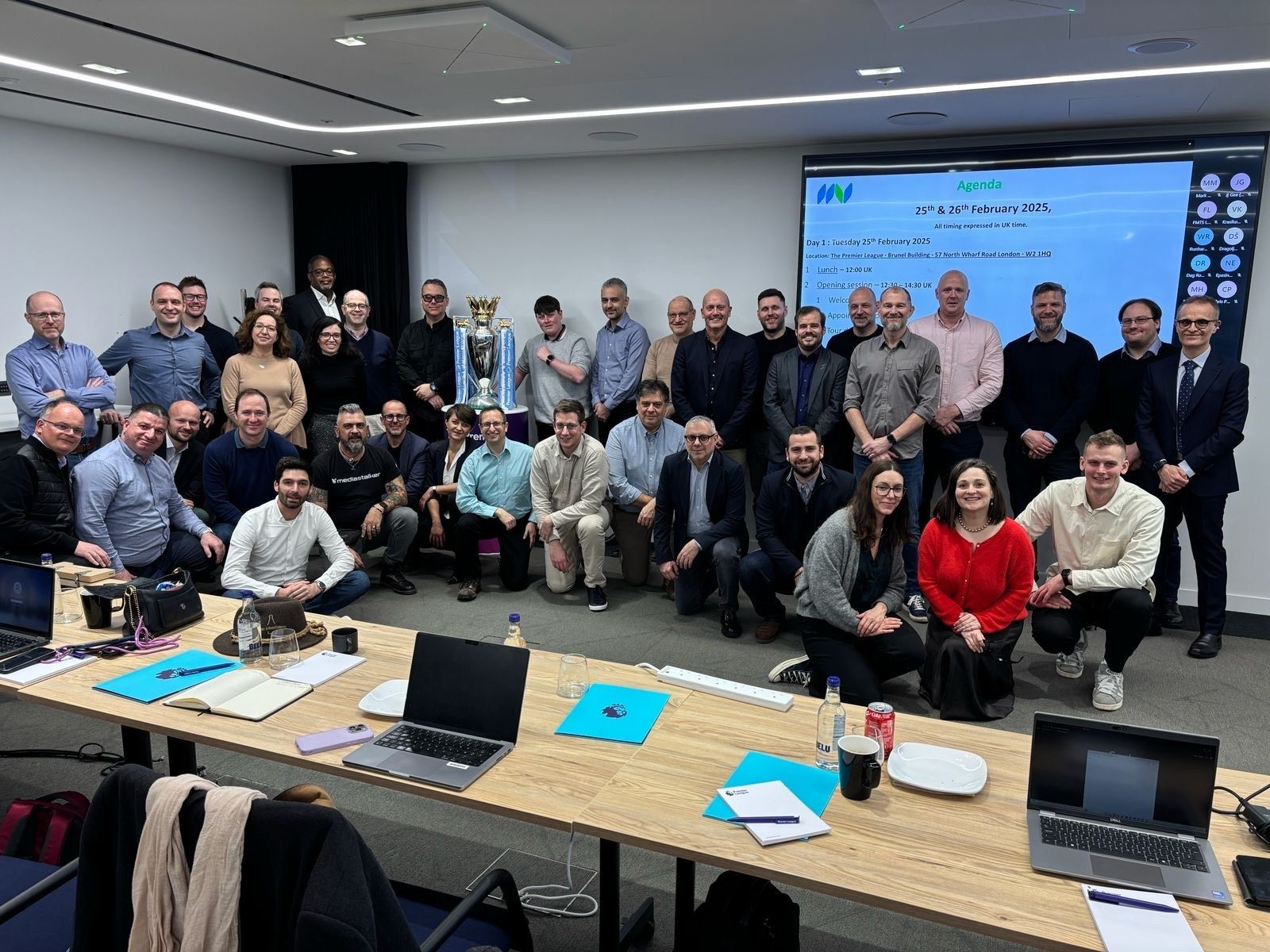 Réunion de travail avec environ quarante participants dans une salle de conférence, autour d'une table de présentation et d'un écran affichant un…